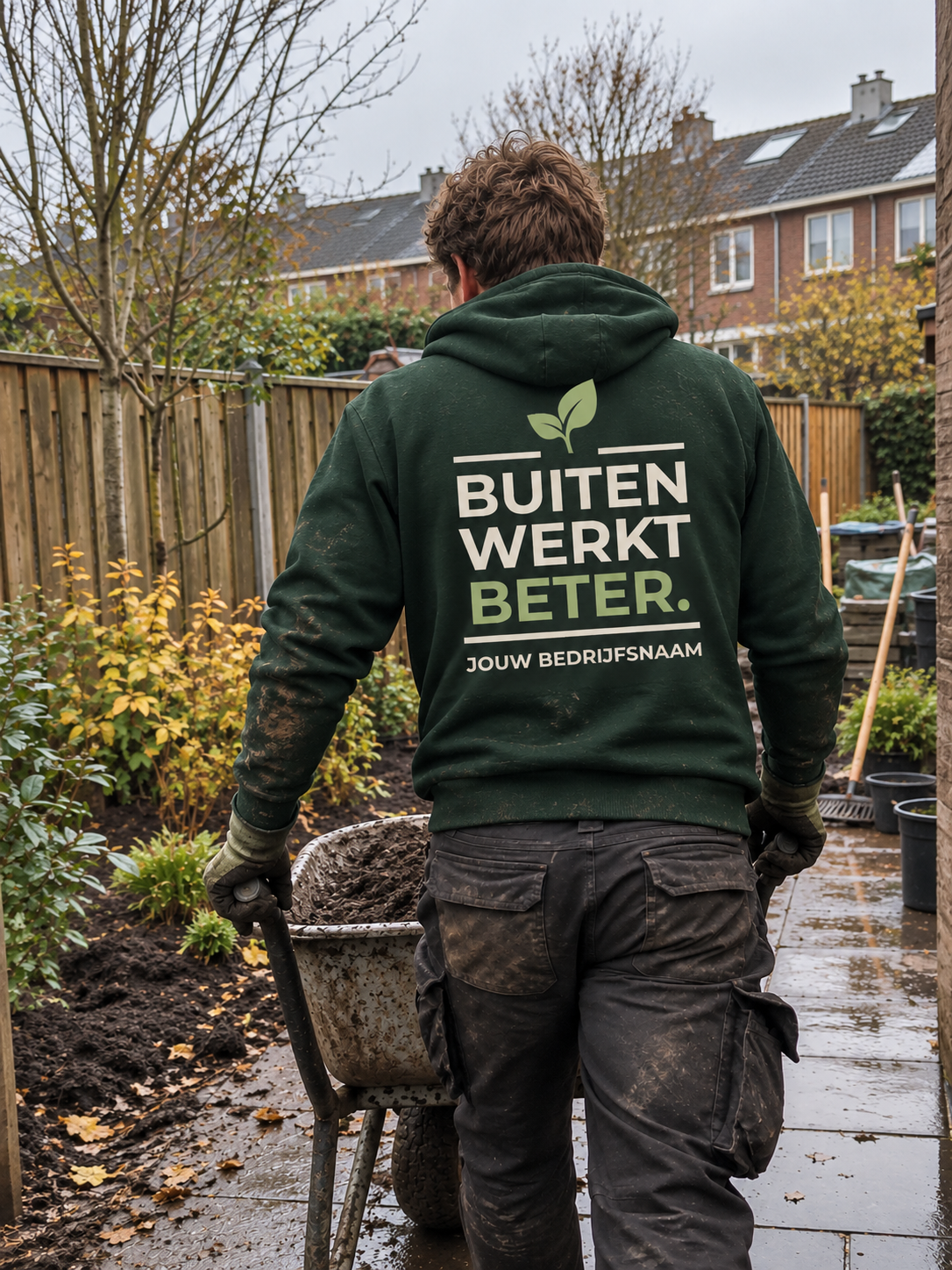 Hovenier in bosgroene hoodie met de tekst "Buiten werkt beter" en bedrijfsnaam op de rug, duwt een kruiwagen door een tuin in de herfst