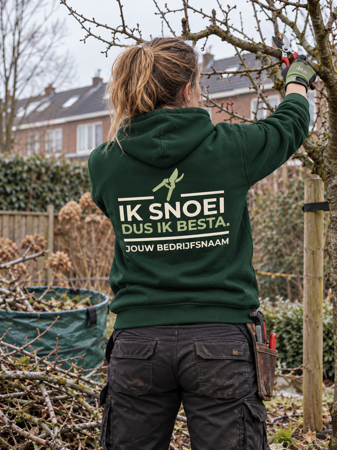 Hovenier in bosgroene hoodie met de tekst "Ik snoei, dus ik besta" en bedrijfsnaam op de rug, snoeit een fruitboom in de winter