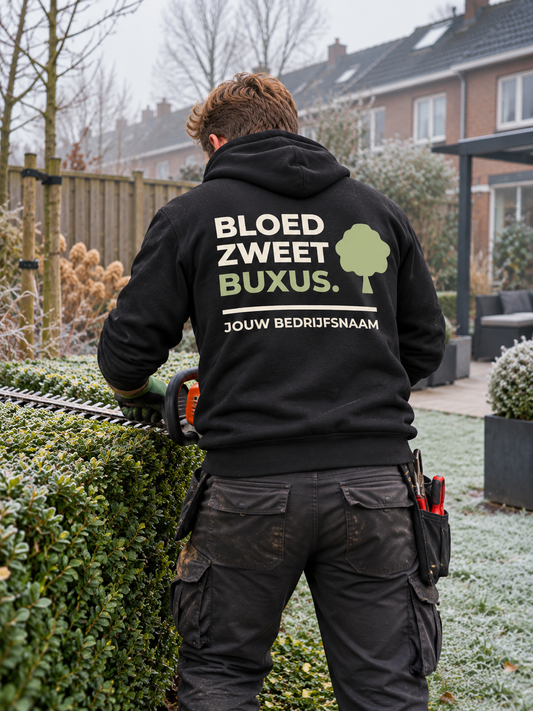 Hovenier in zwarte hoodie met de tekst "Bloed. Zweet. Buxus." en bedrijfsnaam op de rug, snoeit een buxushaag op een koude ochtend