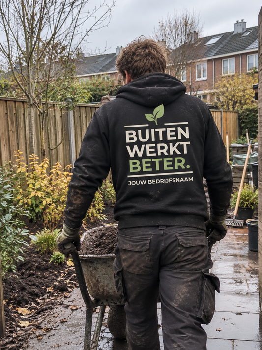 Hovenier in zwart hoodie met de tekst "Buiten werkt beter" en bedrijfsnaam op de rug, duwt een kruiwagen door een tuin in de herfst
